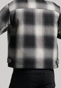 Jacke mit schwarz-weißem Karomuster und leicht strukturierter Optik, ausgestattet mit zwei seitlichen Knöpfen am Saum und einer glatten Textur.