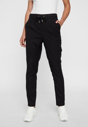 Person wearing slim black drawstring pants and white sneakers, standing with one hand in pocket against plain background.