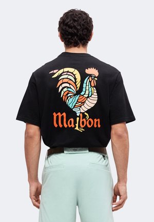 Mand iført en sort T-shirt med en farverig stiliseret hane og ordet "Maibon" trykt på ryggen, kombineret med lys grønne bukser.
