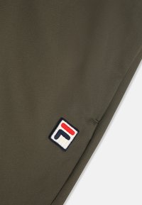 Tejido verde oliva con un patrón hexagonal texturizado, que presenta un logotipo de FILA bordado en rojo, blanco y azul marino en la esquina.