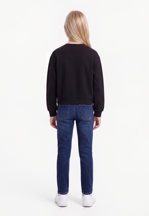 Enfant aux longs cheveux blonds, debout de dos, portant un sweat-shirt noir, un jean bleu et des baskets blanches sur un fond uni.
