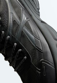 Gros plan sur une chaussure de sport noire montrant une tige en mesh texturé, des superpositions synthétiques noires, des lacets et une semelle en caoutchouc épaisse.