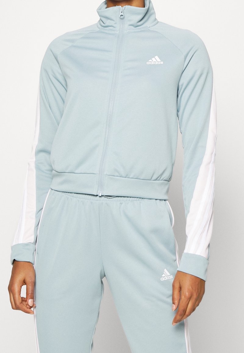 Survêtement Adidas bleu clair avec des bandes blanches sur les manches et le pantalon, veste à zip devant, taille élastique et logos Adidas sur la poitrine et la cuisse.