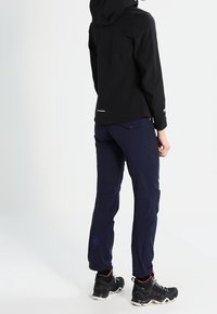 Veste noire ajustée avec capuche, texture lisse ; pantalon bleu marine avec un logo discret, coupe fuselée, et chaussures noires robustes avec des accents rouges.