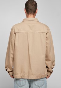 Beige bomullsjacka med avslappnad passform, utrustad med krage, långa ärmar och en platt söm baktill. Bärs över ljusblå jeans.