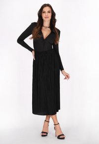 Musta pitkähihainen bodysuit, jossa on v-pääntie ja solmittu etuosa, yhdistettynä laskostettuun mustaan maksihameeseen. Päällä mustat korkokengät.