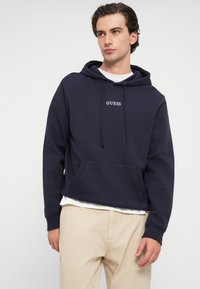 Námornícky modrá mikina s kapucňou vyrobená z mäkkej tkaniny, s prednou kapsou, sťahovacou kapucňou a malým logom "GUESS" na hrudi.