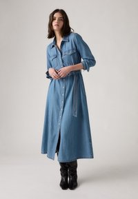 Heled sinine denimpluus-kleit pikkade varrukatega, nööbiga esiosas, kahe rinna taskuga ja sobiva vöödiga, ulatudes põlvani. Mustad põlvikud.