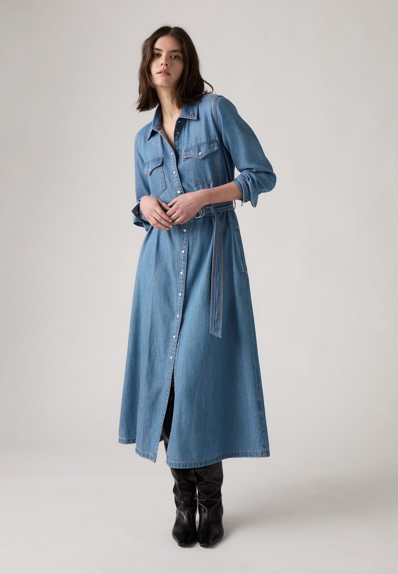 Robe chemise en denim bleu clair avec des manches longues, fermeture à boutons, deux poches poitrine et une ceinture assortie, tombant sous le genou. Bottes noires jusqu'au genou.