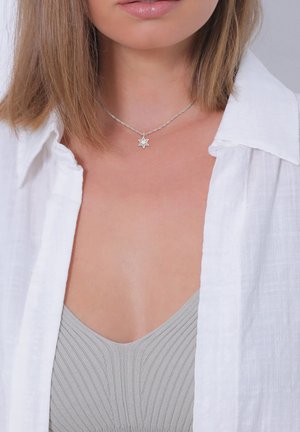 Collana con pendente a stella argentata con catena attorcigliata, indossata sopra una maglia grigia a coste. Lo sfondo presenta un colletto di camicia bianco e texturizzato.