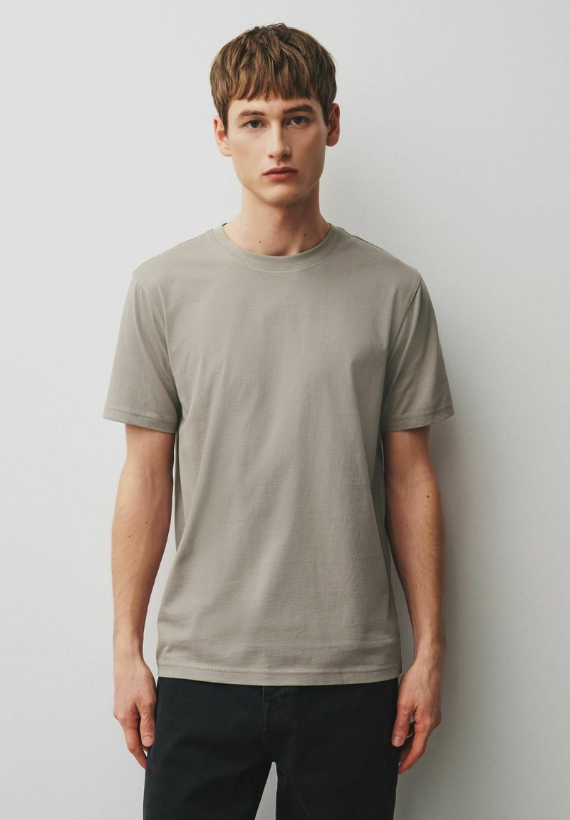 Bărbat tânăr cu păr scurt și castaniu, purtând un tricou simplu cu mânecă scurtă de culoare bej deschis și pantaloni negri, stând în picioare lângă un perete alb.