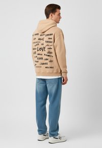 Felpa beige con varie lingue che scrivono "amore" e una tasca a marsupio. Abbinata a jeans blu chiaro e sneakers bianche.