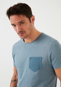 Modrá bavlněná tričko s texturovanou kapsou v mírně tmavším modrém odstínu. Kulatý výstřih a krátké rukávy, hladká textura.