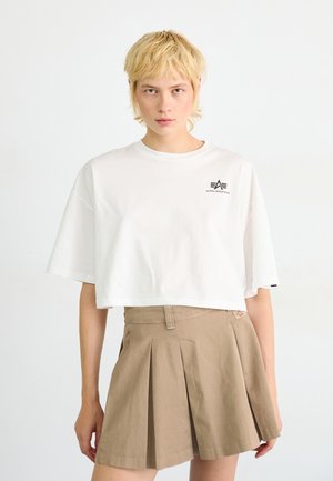 White cropped t-shirt with short sleeves and a small black logo on the chest, paired with a beige pleated skirt featuring visible stitching.