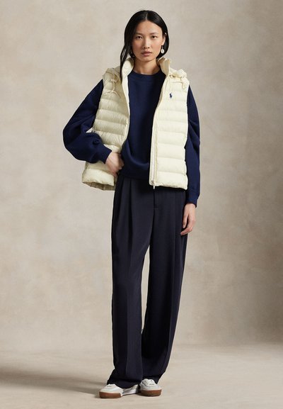 Kvinne med langt svart hår bruker kremfarget dunvest, marineblå genser, vide marineblå bukser og hvite sneakers, står mot beige bakgrunn.