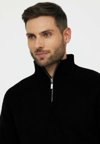 Pull en maille noire avec col montant et fermeture éclair à mi-hauteur. Présente des détails texturés aux épaules et une coupe décontractée.