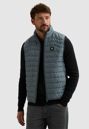 Homme portant un gilet matelassé bleu sur un pull noir, une main dans la poche, regardant légèrement sur le côté.