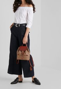 Zaino di stoffa marrone con un flap superiore in pelle e zip, caratterizzato da un motivo con logo. Abbinato a una maglietta bianca off-shoulder e pantaloni scuri.