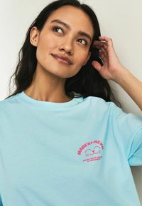 Modra bombažna majica s kratkimi rokavi, na prsih s svetlo rožnatim grafičnim motivom "MANEKI-NEKO" in nasmejanim mačjim dizajnom.