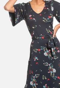 Blommigt mönstrad klänning i mörk marinblått tyg; har en V-ringning, korta vida ärmar, cinchad midja med ett bälte och färgglada blomdetaljer.