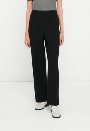 Persona que lleva pantalones negros de talle alto y pierna recta, camiseta gris de manga corta y zapatos blancos sin cordones, de pie frente a un fondo blanco.