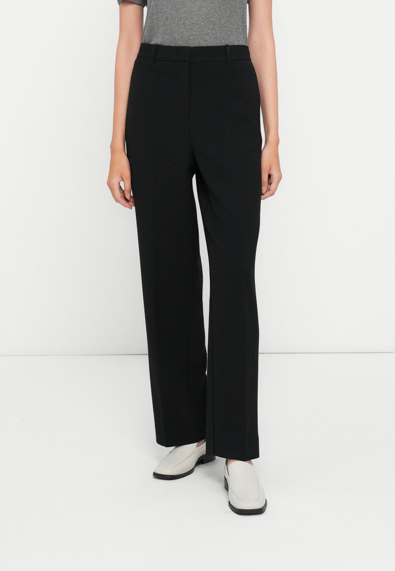 Personne portant un pantalon noir taille haute droit, un haut gris à manches courtes et des chaussures blanches à enfiler, debout devant un fond blanc.