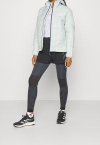 Veste matelassée bleu clair avec capuche, associée à un short de sport noir et des leggings texturés. Chaussures de course noires et détails du logo visibles.