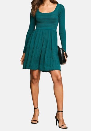 Femme portant une robe en maille bleu sarcelle à manches longues avec un col et un ourlet festonnés, des sandales à talons noires et tenant un sac à main noir.