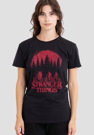 Junge Frau trägt ein schwarzes Stranger Things T-Shirt mit rotem Aufdruck von Kindern, die mit Fahrrädern fahren, und Bäumen vor einem roten Sonnenhintergrund.