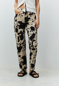 Pantalon beige clair avec de grandes taches noires, coupe droite, cinq poches, associé à des sandales plates noires.