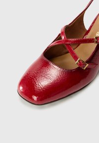 Chaussure en cuir verni rouge avec une finition brillante, dotée d'un bout ouvert, de brides croisées et d'un petit talon carré.