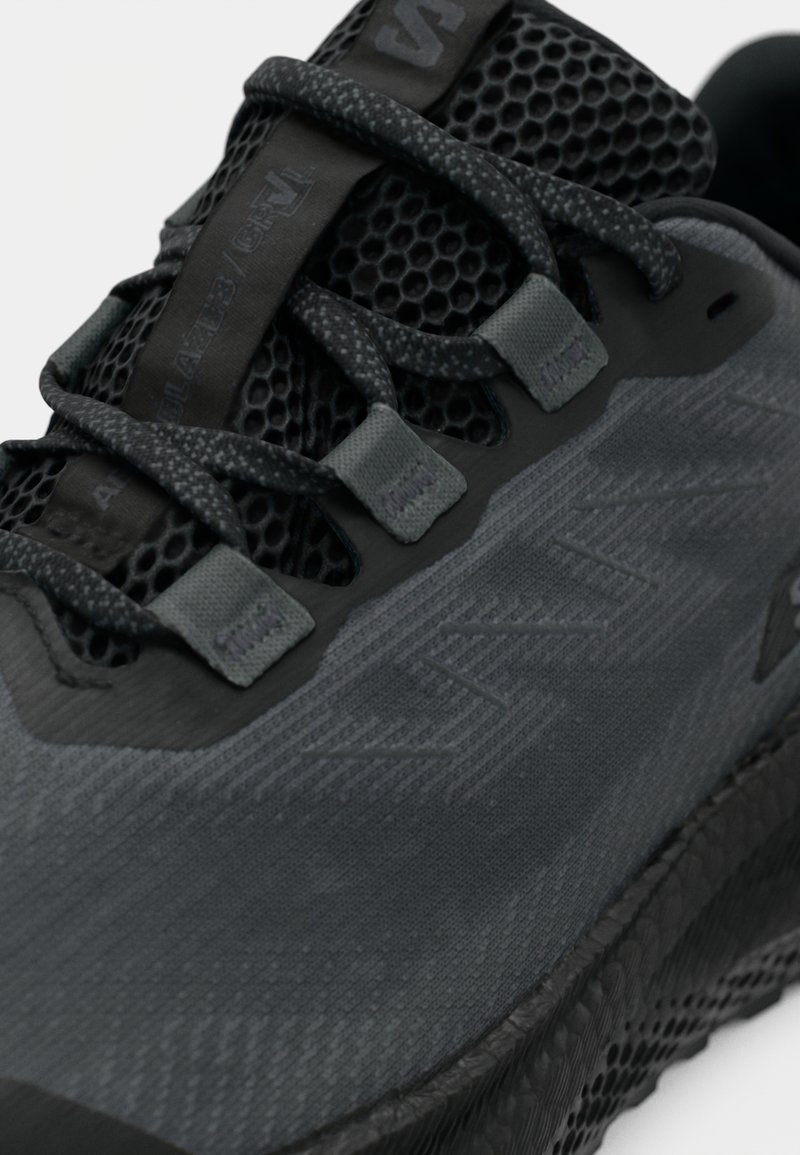 Dettaglio ravvicinato di una scarpa sportiva nera e grigio scuro con tomaia in rete texturizzata, lacci a motivo e dettaglio della suola robusta.