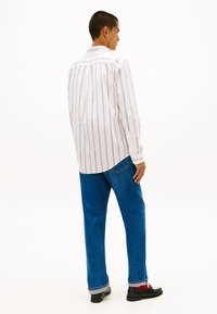 Λευκό πουκάμισο με λεπτές κόκκινες ρίγες, κομμένο μέσα σε μπλε τζιν. Το πουκάμισο έχει κλασικό γιακά και μακριά μανίκια, με στρογγυλεμένο τελείωμα.