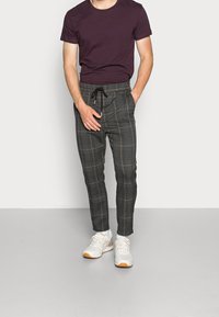Homme portant une chemise à manches courtes violet foncé, un pantalon à carreaux gris avec cordon de serrage, et des baskets blanches, debout devant un fond uni.