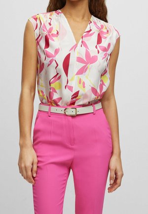 Femme portant un chemisier sans manches blanc avec un imprimé floral rose et jaune, rentré dans un pantalon taille haute rose vif avec une ceinture blanche.