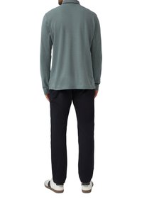 Homme portant une chemise à manches longues gris-vert, un pantalon noir et des baskets blanches, tourné le dos contre un fond blanc.