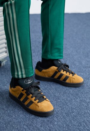 Persona que lleva pantalones deportivos verdes de adidas y zapatillas adidas mostaza con rayas negras, de pie sobre una alfombra azul.