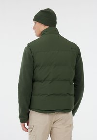 Man in een donkergroene beanie, bijpassende gewatteerde vest en fleece jas, met beige cargobroek, met zijn rug naar de camera gekeerd op een witte achtergrond.