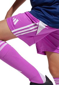 Roze sportshorts met witte accenten, met aan elke kant drie witte strepen. Paarse, geribbelde kniehoge sokken en zwarte schoenen zichtbaar.