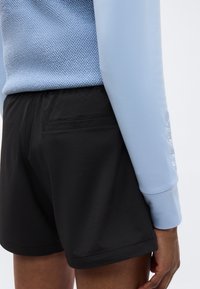 Άτομο που φοράει μαύρο σορτς με πίσω τσέπη και ανοιχτό μπλε μακρυμάνικο τοπ με υφή, εμφανιζόμενο από τη μέση και κάτω.