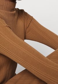 Hnědý žebrovaný rolák s přiléhavým střihem a dlouhými rukávy. Textura má vertikální žebrování pro zvýraznění detailů.