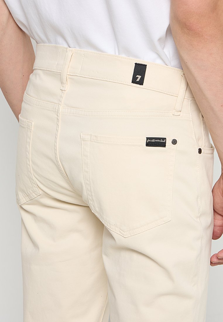 Beige katoenen broek met een rechte snit, twee achterzakken en een zwart merketiket op de tailleband. De stof lijkt glad en duurzaam.