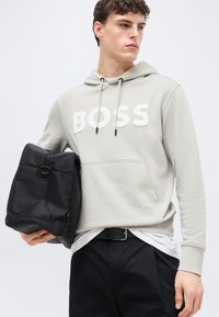 Jeune homme portant un sweat à capuche BOSS gris clair, tenant un sac noir plié, vêtu d'un pantalon noir et d'une chemise blanche sous le sweat.