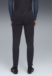 Pantaloni sportivi negri dintr-un material ușor, cu o croială slim, având fermoare laterale la gleznă și asortați cu ghete negre.
