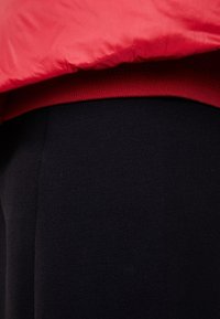 Rød polstret jakke over en rød sweater, kombineret med sorte bukser i stof. Glatte teksturer og kontrasterende farver skaber et lagdelt udseende.