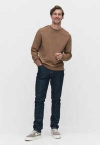 Bruine sweatshirt met een ronde hals, gecombineerd met donkerblauwe jeans en lichtgrijze sneakers. Model staat met handen in de zakken.