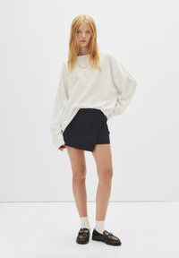 Top blanc oversized à manches longues, jupe enveloppante noir à rayures, chaussures noires à boucles, et chaussettes blanches à la cheville sur fond uni.