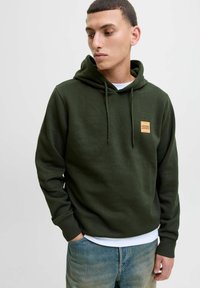 Dark green hooded sweatshirt made of soft cotton, featuring a front label patch, adjustable drawstrings, and ribbed cuffs. Worn with blue jeans.