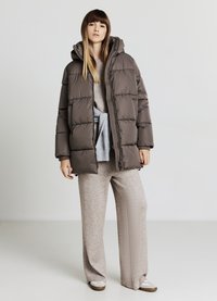 Brown puffer jacket with a hood, featuring a quilted design. Worn over a light gray sweater and beige wide-leg pants. Casual footwear visible.