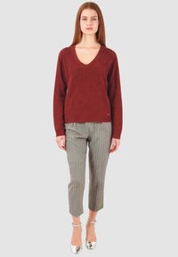 Maglione a V in maglia borgogna, pantaloni sartoriali grigi con righe sottili e scarpe con tacco argento su uno sfondo semplice e neutro.
